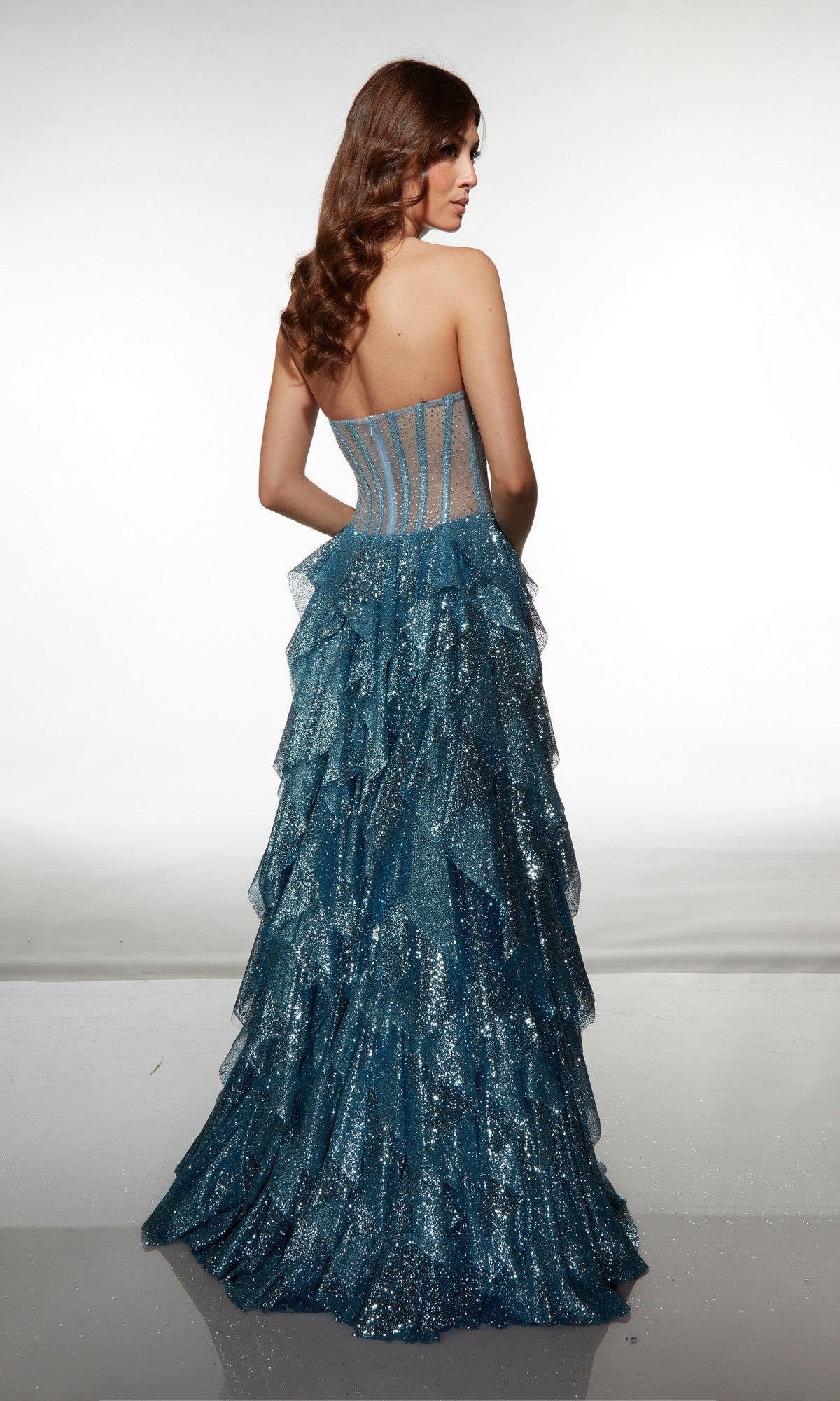 A woman with long brown hair wears the Alyce 62081, a strapless blue glitter corset dress with a sheer back and layered ruffled skirt, standing against a plain white background.